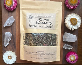 May include: A kraft paper pouch of Maine Blueberry herbal tea blend with a clear window, surrounded by dried flowers and quartz crystals. The label reads "Royal Pine Apothecary" and lists ingredients. The tea is displayed on a wooden cutting board.