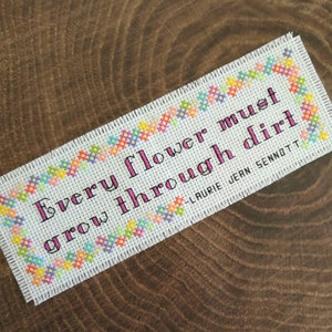 May include: A white cross-stitch bookmark with a colorful floral border. The bookmark features the quote "Every flower must grow through dirt" attributed to Laurie Jean Sennott.