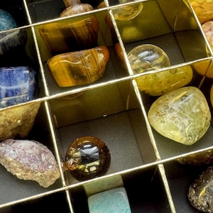 Könnte beinhalten: Eine Auswahl an polierten Edelsteinen in einer unterteilten Box. Die Steine umfassen Tigerauge, Quarz und andere farbenfrohe Mineralien. Die Box hat goldfarbene Trennwände und einen schwarzen Innenraum, wodurch eine visuell organisierte Präsentation natürlicher Schätze entsteht.