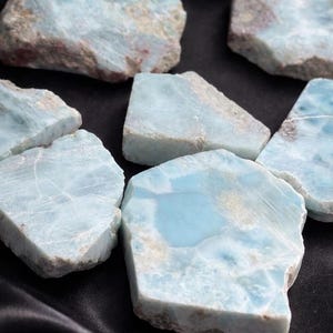 May include: A collection of raw larimar stones, showcasing a pale blue hue with white and brown marbling. The stones are irregularly shaped and vary in size, resting on a black, textured surface.