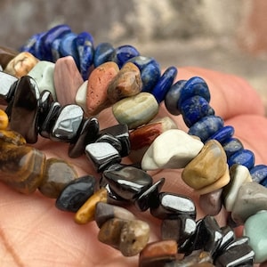 May include: Close-up of several strands of gemstone chip bracelets. The bracelets feature a variety of colours, including blue, brown, black, and white. The gemstones are irregularly shaped and appear to be strung together.