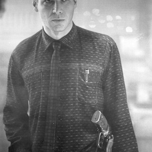 Puede incluir: Imagen en blanco y negro de un hombre con una camisa estampada y corbata. Un bol&iacute;grafo est&aacute; en el bolsillo de la camisa. Una pistola est&aacute; en una funda en la cadera. El hombre sostiene un vaso de l&iacute;quido.
