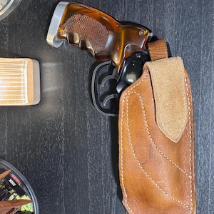 Puede incluir: Una pistola con empu&ntilde;adura marr&oacute;n y ca&ntilde;&oacute;n negro est&aacute; parcialmente enfundada en una funda de cuero marr&oacute;n. La funda tiene una solapa marr&oacute;n claro y est&aacute; cosida con hilo blanco. El arma descansa sobre una superficie oscura.
