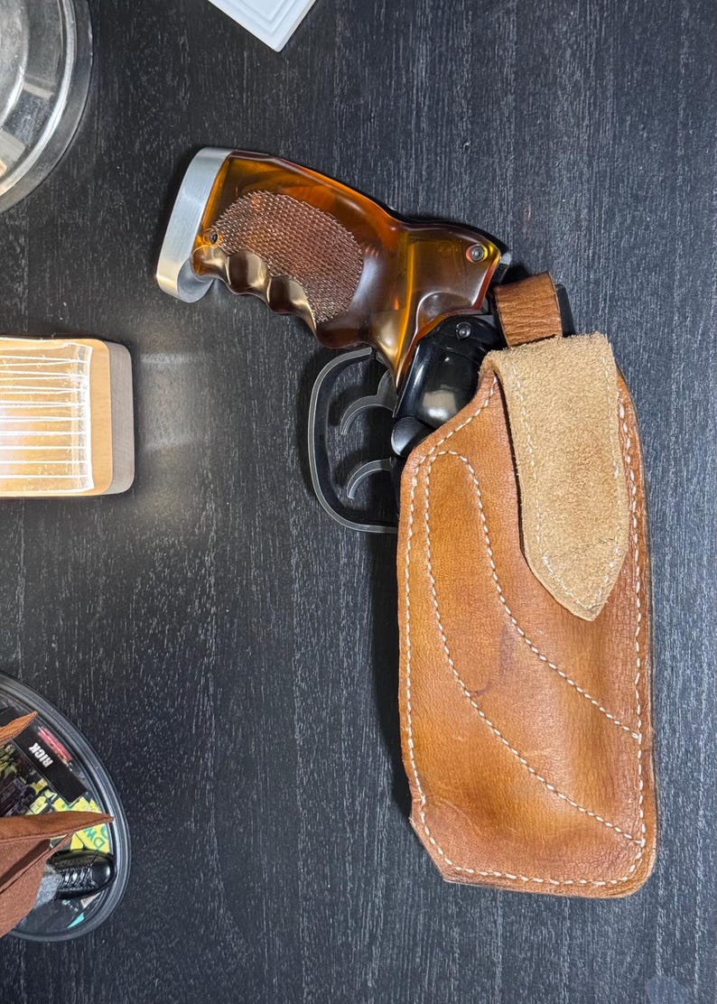 Puede incluir: Una pistola con empu&ntilde;adura marr&oacute;n y ca&ntilde;&oacute;n negro est&aacute; parcialmente enfundada en una funda de cuero marr&oacute;n. La funda tiene una solapa marr&oacute;n claro y est&aacute; cosida con hilo blanco. El arma descansa sobre una superficie oscura.