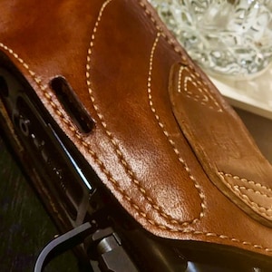 Puede incluir: Primer plano de una funda de cuero marr&oacute;n con costuras blancas, que oculta parcialmente una pistola negra. La funda tiene una superficie texturizada y una solapa. Un vaso transparente con un dise&ntilde;o texturizado est&aacute; en el fondo.