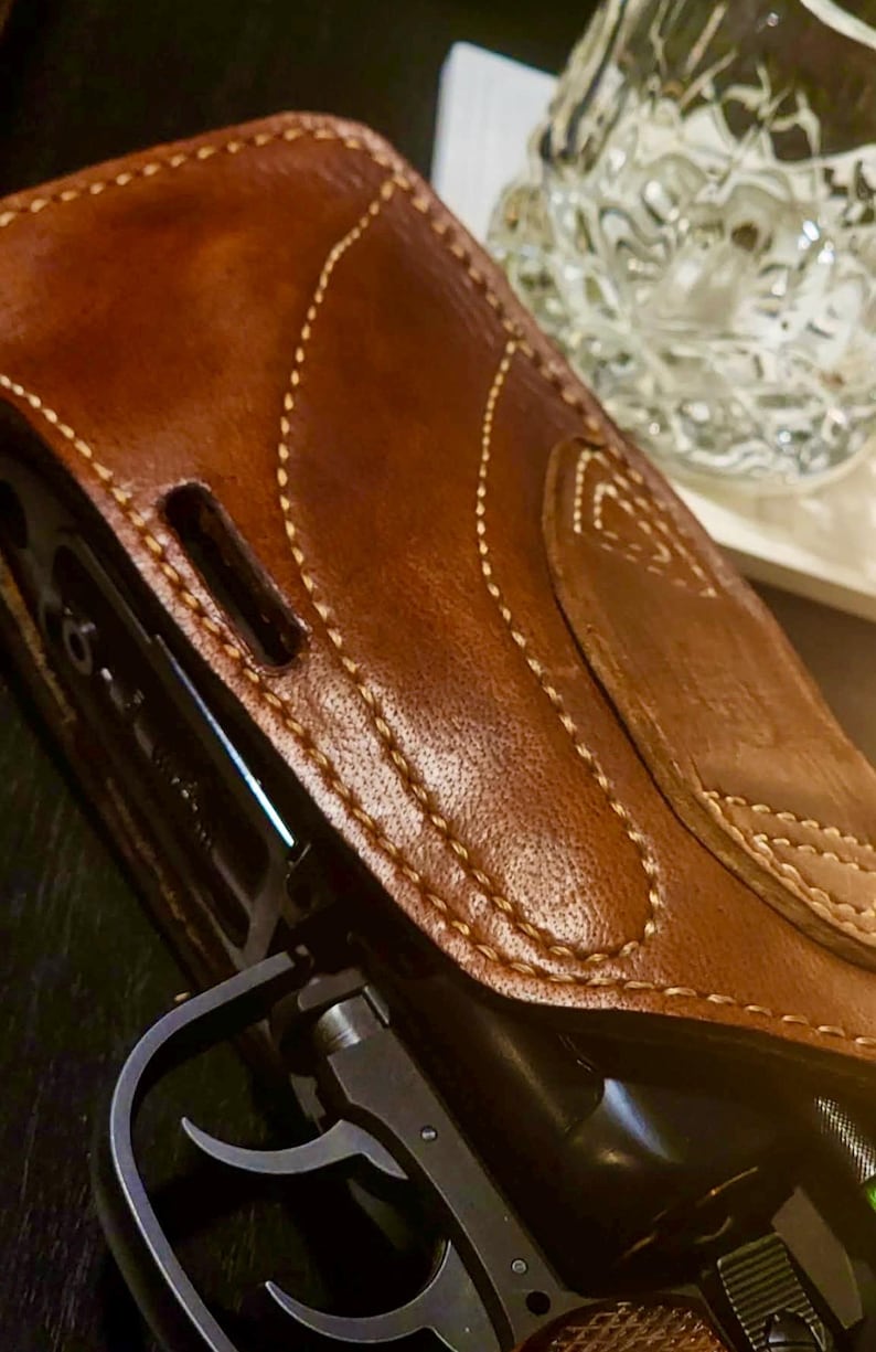 Puede incluir: Primer plano de una funda de cuero marr&oacute;n con costuras blancas, que oculta parcialmente una pistola negra. La funda tiene una superficie texturizada y una solapa. Un vaso transparente con un dise&ntilde;o texturizado est&aacute; en el fondo.