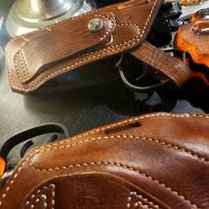 Puede incluir: Primer plano de dos fundas de pistola de cuero marr&oacute;n con costuras blancas. Una funda est&aacute; abierta, revelando un arma. Las fundas tienen un dise&ntilde;o cl&aacute;sico con cierre de bot&oacute;n a presi&oacute;n y probablemente est&aacute;n hechas a mano.