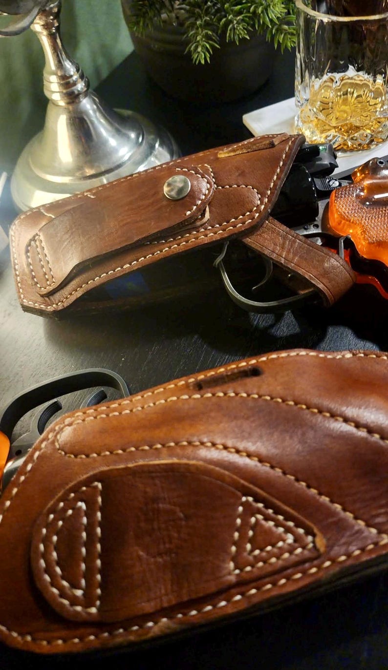 Puede incluir: Primer plano de dos fundas de pistola de cuero marr&oacute;n con costuras blancas. Una funda est&aacute; abierta, revelando un arma. Las fundas tienen un dise&ntilde;o cl&aacute;sico con cierre de bot&oacute;n a presi&oacute;n y probablemente est&aacute;n hechas a mano.