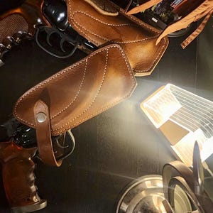 Puede incluir: Una funda de cuero marr&oacute;n con costuras blancas y una correa, dise&ntilde;ada para sujetar un rev&oacute;lver. La imagen tambi&eacute;n muestra un rev&oacute;lver con empu&ntilde;adura marr&oacute;n y ca&ntilde;&oacute;n negro. Los art&iacute;culos est&aacute;n sobre una superficie oscura.