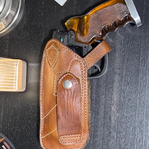 Puede incluir: Una funda de cuero marr&oacute;n con una correa a juego y un cierre de bot&oacute;n plateado. Una pistola con una empu&ntilde;adura marr&oacute;n y plateada es parcialmente visible dentro de la funda. La imagen est&aacute; tomada desde una perspectiva cenital.