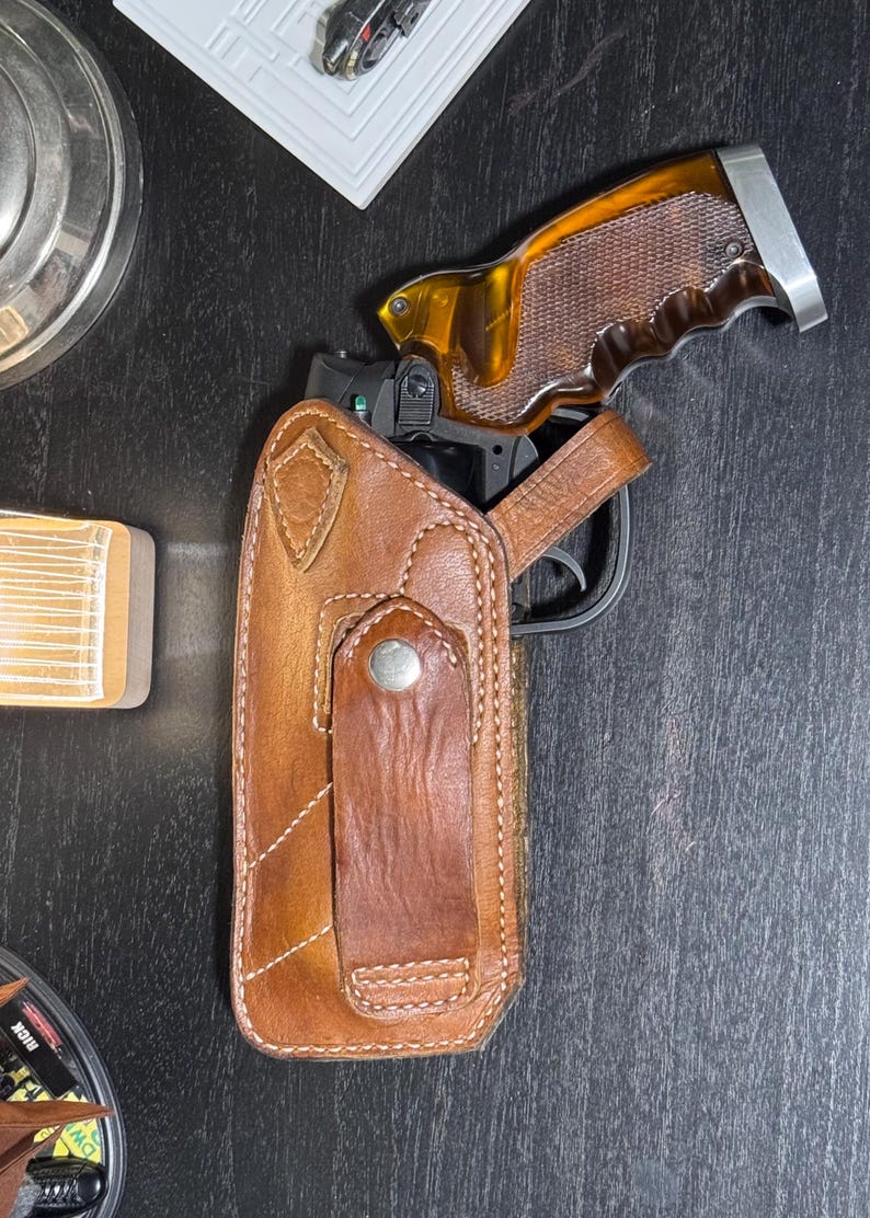 Puede incluir: Una funda de cuero marr&oacute;n con una correa a juego y un cierre de bot&oacute;n plateado. Una pistola con una empu&ntilde;adura marr&oacute;n y plateada es parcialmente visible dentro de la funda. La imagen est&aacute; tomada desde una perspectiva cenital.