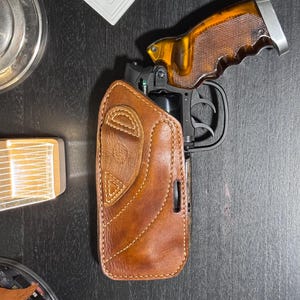 May include: A brown leather holster with a stitched design, holding a black handgun with a brown and silver grip. The holster is on a dark surface.