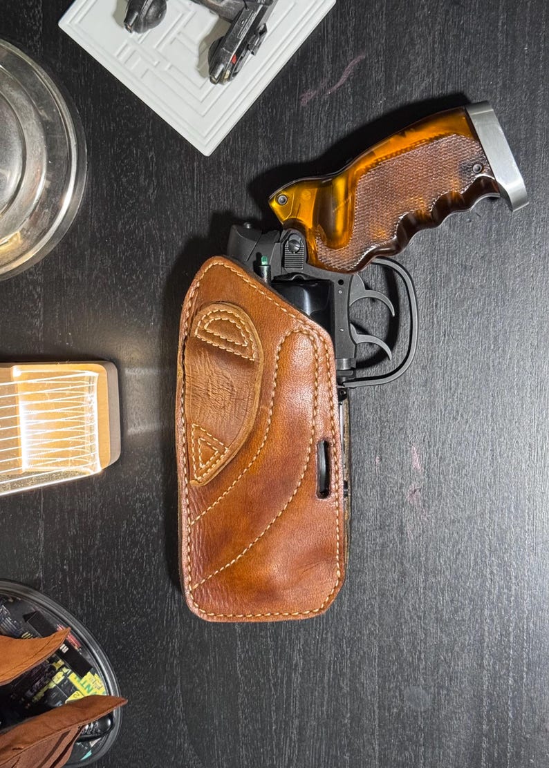 Puede incluir: Una funda de cuero marr&oacute;n con un dise&ntilde;o cosido, que sostiene una pistola negra con una empu&ntilde;adura marr&oacute;n y plateada. La funda est&aacute; sobre una superficie oscura.