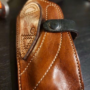 Puede incluir: Una funda de cuero artesanal en tonos marrones, con costuras intrincadas y un cierre de correa negro seguro. La funda tiene una superficie texturizada y un dise&ntilde;o cosido decorativo en la solapa.
