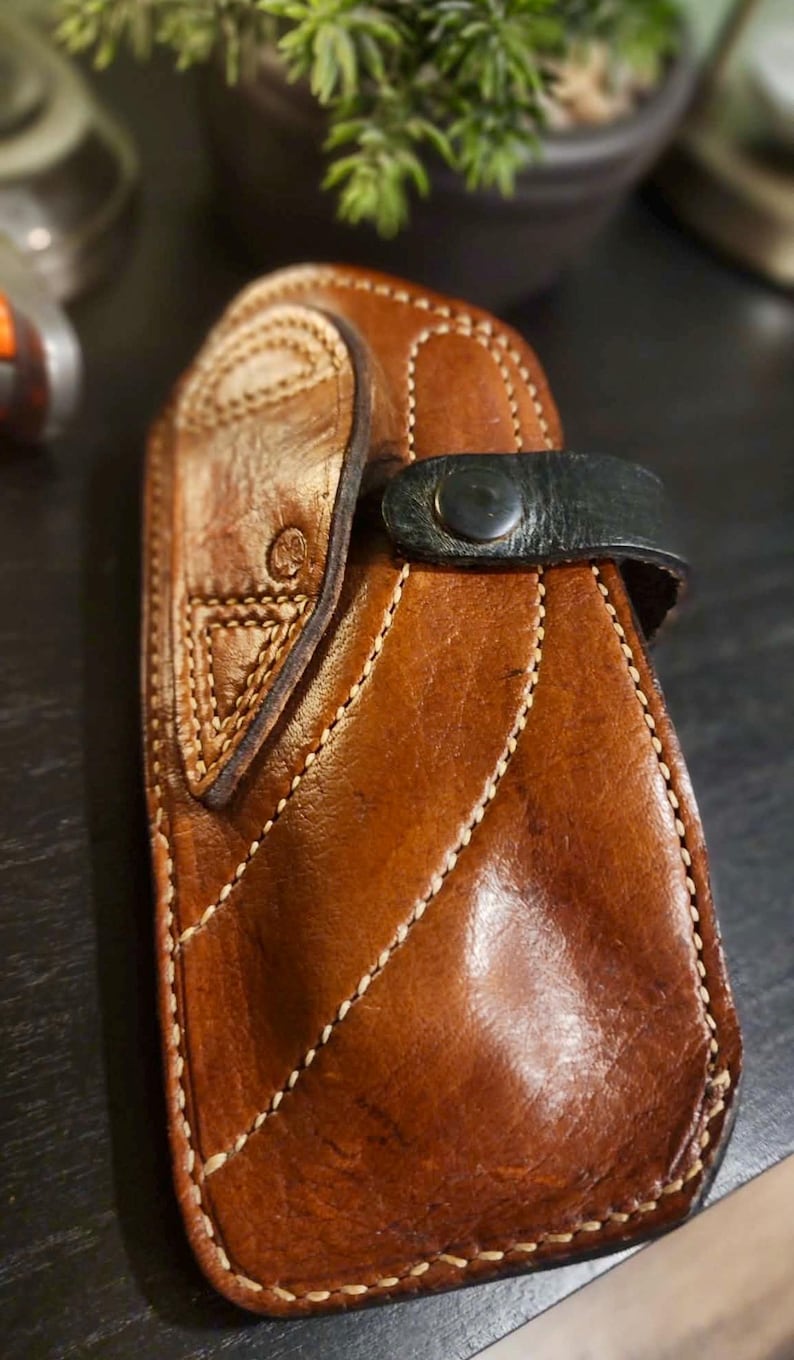 Puede incluir: Una funda de cuero artesanal en tonos marrones, con costuras intrincadas y un cierre de correa negro seguro. La funda tiene una superficie texturizada y un dise&ntilde;o cosido decorativo en la solapa.