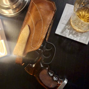 Puede incluir: Una funda de cuero marr&oacute;n con costuras blancas sostiene una pistola negra con empu&ntilde;adura de madera. El arma es parcialmente visible, apoyada sobre una superficie oscura. Un vaso de l&iacute;quido &aacute;mbar est&aacute; cerca.