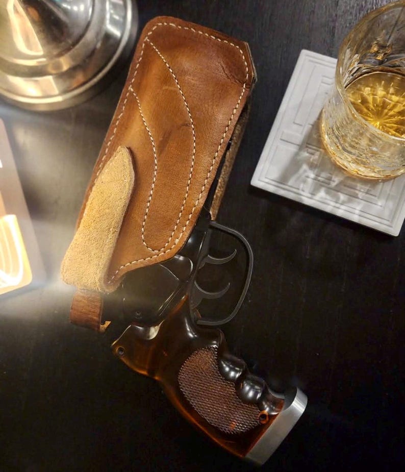 Puede incluir: Una funda de cuero marr&oacute;n con costuras blancas sostiene una pistola negra con empu&ntilde;adura de madera. El arma es parcialmente visible, apoyada sobre una superficie oscura. Un vaso de l&iacute;quido &aacute;mbar est&aacute; cerca.