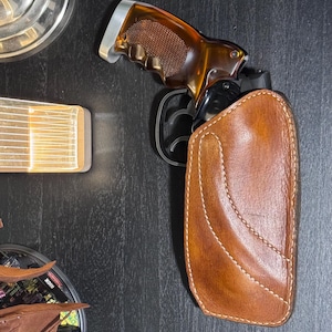 Puede incluir: Un rev&oacute;lver marr&oacute;n con una empu&ntilde;adura marr&oacute;n y plateada est&aacute; parcialmente insertado en una funda de cuero marr&oacute;n. La funda tiene costuras blancas. El rev&oacute;lver es negro y marr&oacute;n.