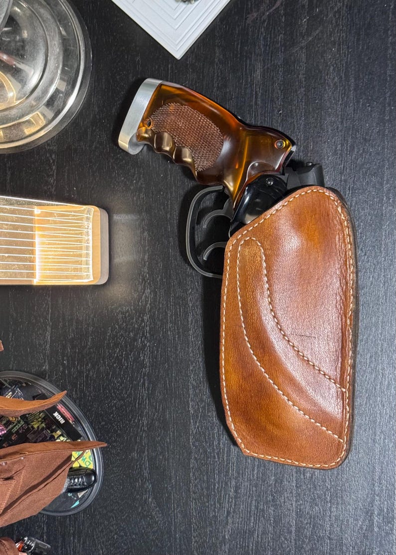 Puede incluir: Un rev&oacute;lver marr&oacute;n con una empu&ntilde;adura marr&oacute;n y plateada est&aacute; parcialmente insertado en una funda de cuero marr&oacute;n. La funda tiene costuras blancas. El rev&oacute;lver es negro y marr&oacute;n.