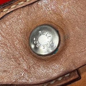 Puede incluir: Primer plano de un art&iacute;culo de cuero marr&oacute;n con un bot&oacute;n de metal plateado. El bot&oacute;n tiene grabadas las palabras "PULL THE DOT". El cuero tiene costuras visibles y una superficie texturizada.