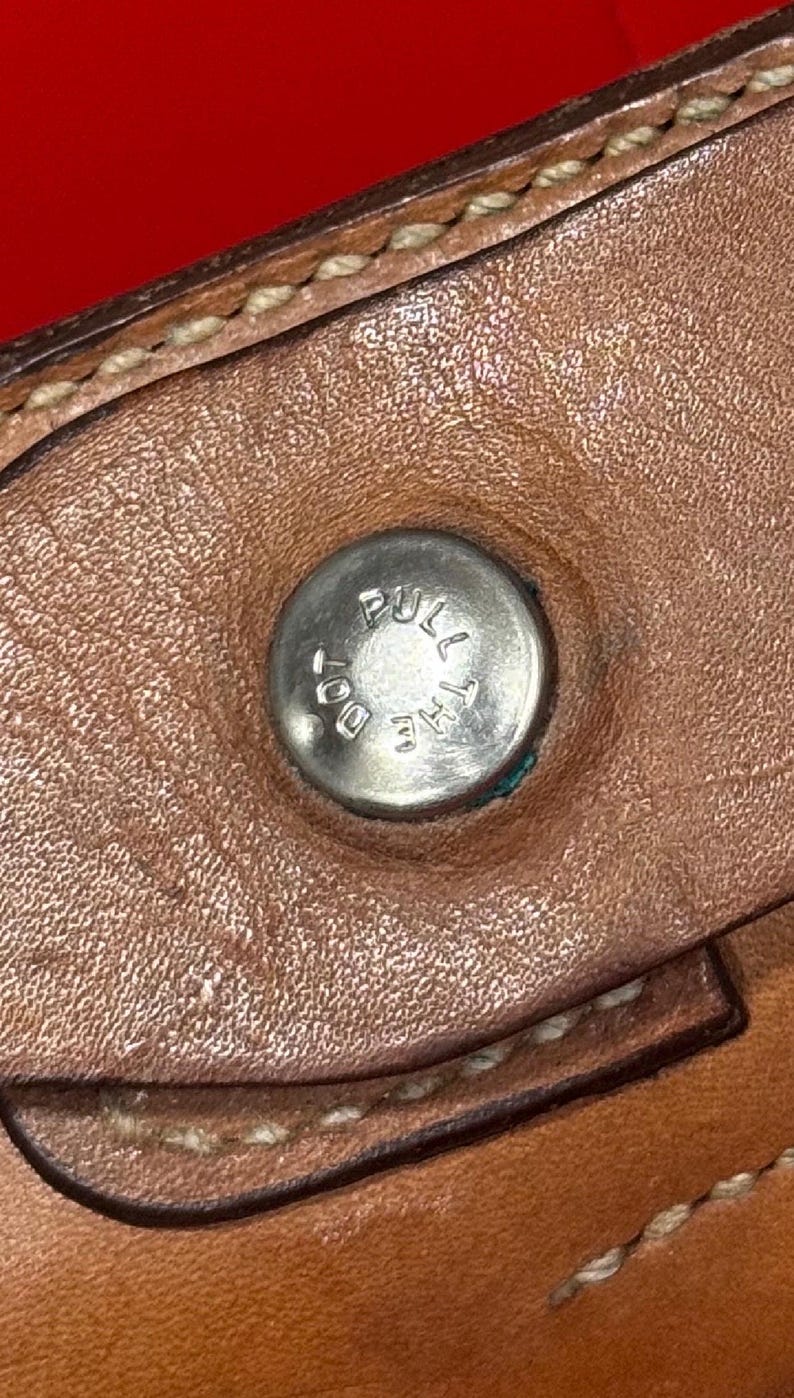 Puede incluir: Primer plano de un art&iacute;culo de cuero marr&oacute;n con un bot&oacute;n de metal plateado. El bot&oacute;n tiene grabadas las palabras "PULL THE DOT". El cuero tiene costuras visibles y una superficie texturizada.