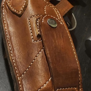 Puede incluir: Una funda de cuero marr&oacute;n con costuras blancas y cierre de bot&oacute;n a presi&oacute;n. La funda parece ser para una pistola, con una parte del arma visible. El cuero tiene un aspecto desgastado.