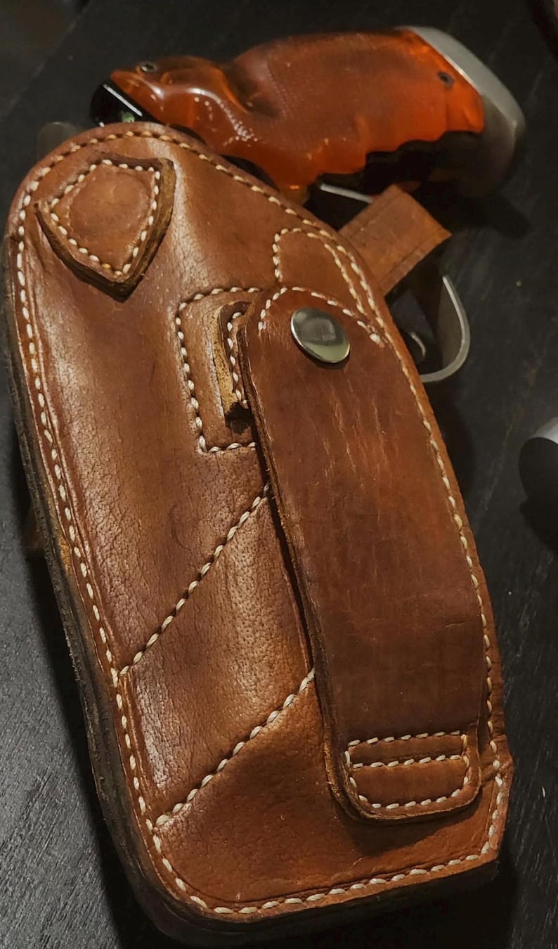 Puede incluir: Una funda de cuero marr&oacute;n con costuras blancas y cierre de bot&oacute;n a presi&oacute;n. La funda parece ser para una pistola, con una parte del arma visible. El cuero tiene un aspecto desgastado.