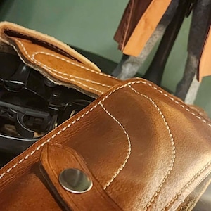 Puede incluir: Primer plano de una funda de cuero marr&oacute;n con costuras blancas y un broche plateado. La funda est&aacute; dise&ntilde;ada para guardar un arma. Una figura con un abrigo marr&oacute;n y una pistola est&aacute; en el fondo.