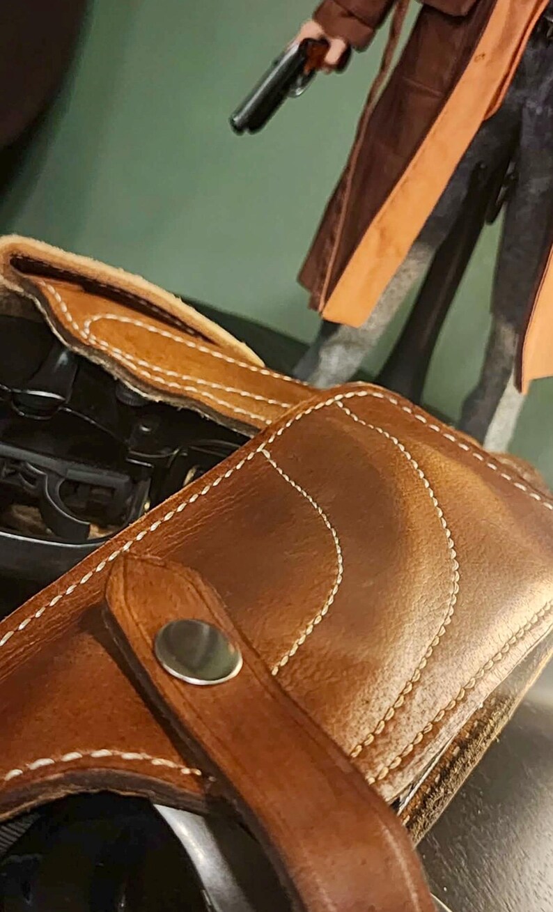 Puede incluir: Primer plano de una funda de cuero marr&oacute;n con costuras blancas y un broche plateado. La funda est&aacute; dise&ntilde;ada para guardar un arma. Una figura con un abrigo marr&oacute;n y una pistola est&aacute; en el fondo.