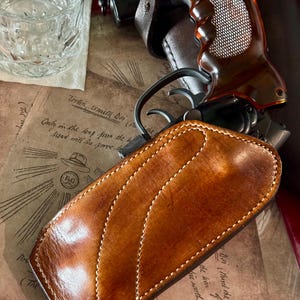 Puede incluir: Primer plano de una funda de cuero marr&oacute;n con costuras blancas, una pistola con empu&ntilde;adura marr&oacute;n y un vaso de agua. La funda es el foco principal, con un dise&ntilde;o curvo. El fondo incluye papel de estilo vintage con texto manuscrito.