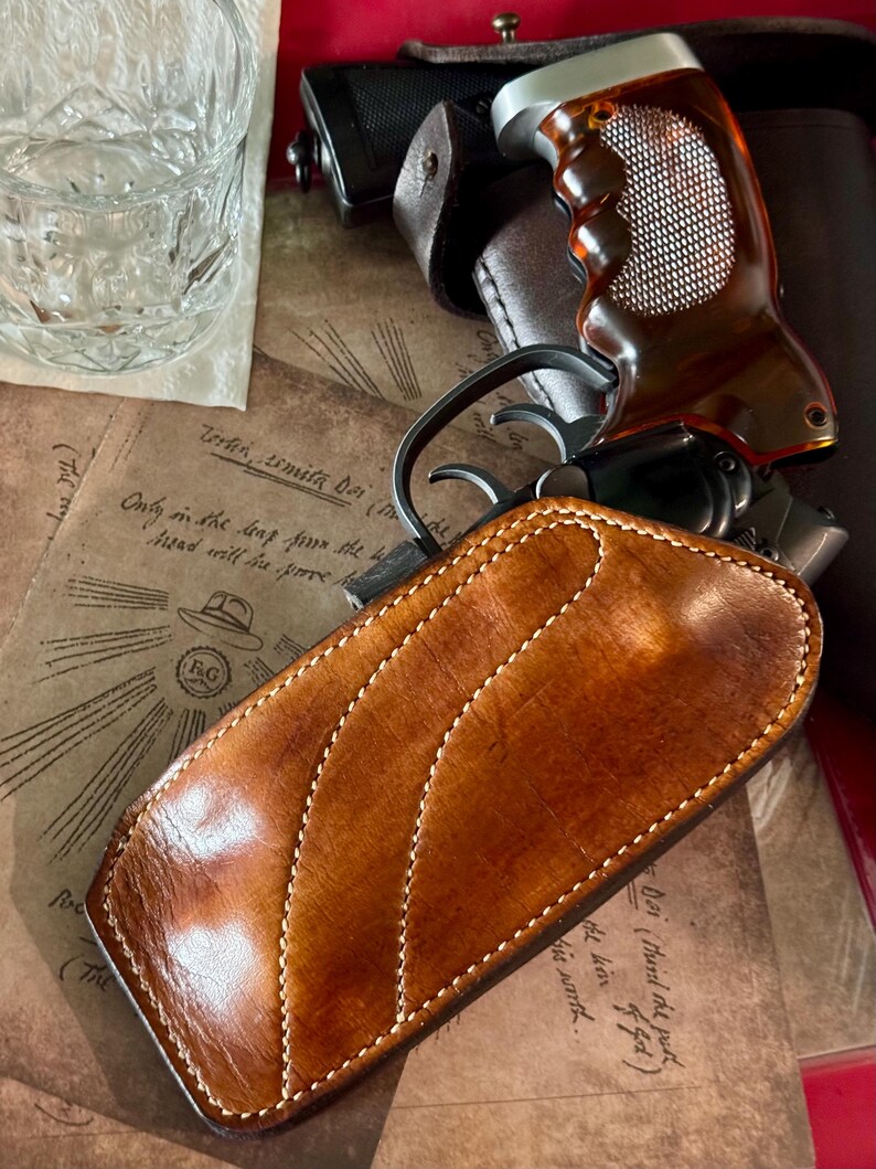 Puede incluir: Primer plano de una funda de cuero marr&oacute;n con costuras blancas, una pistola con empu&ntilde;adura marr&oacute;n y un vaso de agua. La funda es el foco principal, con un dise&ntilde;o curvo. El fondo incluye papel de estilo vintage con texto manuscrito.