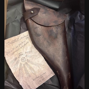 May include: A dark brown leather holster with a flap closure and a metal stud, accompanied by a piece of aged parchment paper featuring handwritten text and a drawing of a hat with rays of light, all resting on black tissue paper.