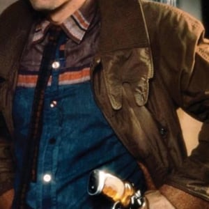 Puede incluir: Un hombre con una chaqueta de cuero marr&oacute;n, camisa azul y pantalones oscuros, con una pistola en una funda. La chaqueta tiene un cuello ancho y la camisa tiene un estampado de rayas. La escena est&aacute; tenuemente iluminada.