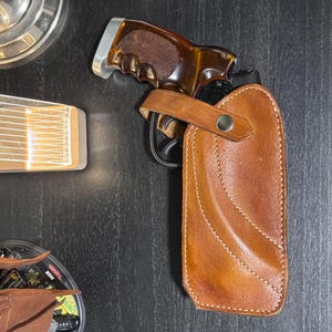 Puede incluir: Una pistola con empu&ntilde;adura marr&oacute;n est&aacute; parcialmente insertada en una funda de cuero marr&oacute;n. La funda tiene un dise&ntilde;o cosido y una correa con un bot&oacute;n plateado. El fondo es una superficie de madera oscura.