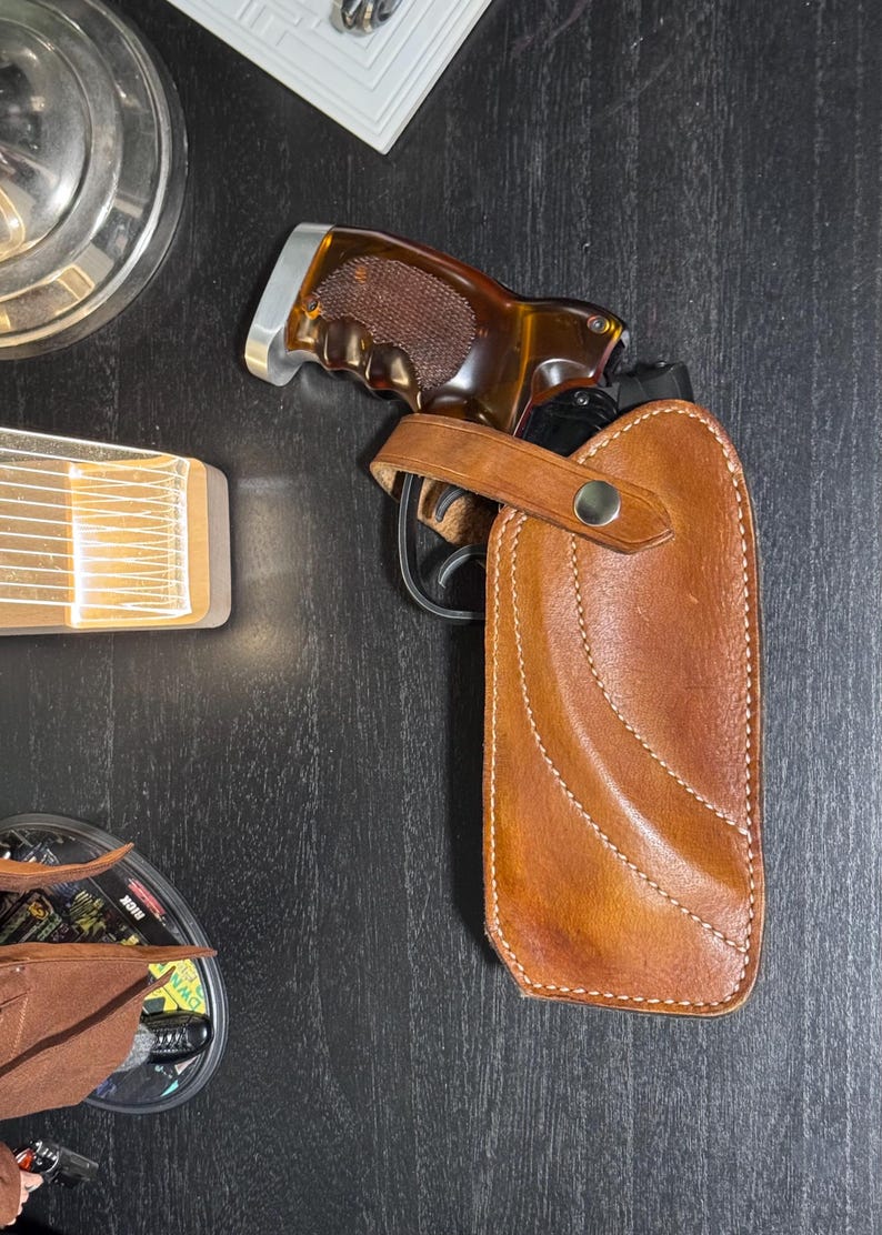 Puede incluir: Una pistola con empu&ntilde;adura marr&oacute;n est&aacute; parcialmente insertada en una funda de cuero marr&oacute;n. La funda tiene un dise&ntilde;o cosido y una correa con un bot&oacute;n plateado. El fondo es una superficie de madera oscura.