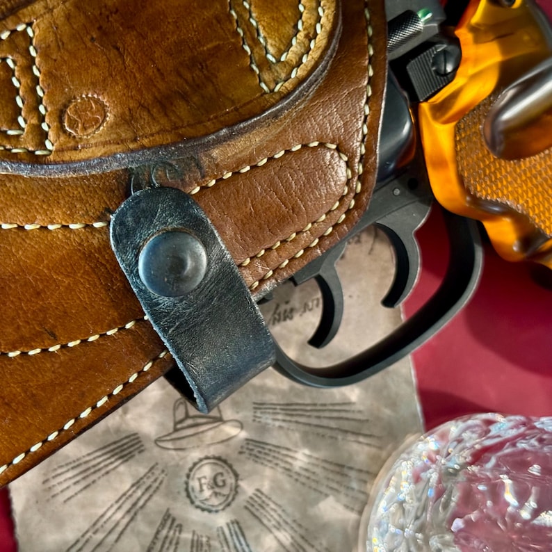 Puede incluir: Primer plano de una funda de cuero marr&oacute;n con costuras blancas y una correa negra. Un rev&oacute;lver negro con empu&ntilde;adura naranja es parcialmente visible. Un papel vintage con las iniciales "F.G" y una ilustraci&oacute;n de sombrero.