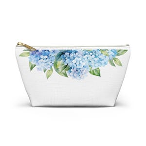 Puede incluir: Bolsa de cosméticos blanca con cremallera dorada y un diseño de acuarela de flores de hortensia azules y hojas verdes en la parte superior. La bolsa está hecha de un material similar a la lona.