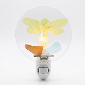 May include: A white plug-in night light featuring a clear circular glass panel. The glass is decorated with a yellow dragonfly silhouette, a smooth amber-colored piece of sea glass, and a light blue piece of sea glass near the base.