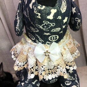May include: A denim dog toy with a white lace collar and a white bow. The toy has a black nose and is covered in white stars and bone patterns.