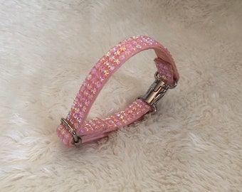 Puede incluir: Un collar para perro rosa con pedrería iridiscente. El collar tiene una hebilla plateada y está hecho de un material suave y flexible.