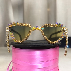 Op de afbeelding: Gouden cat-eye zonnebril met iriserende strass steentjes en een kristal ketting detail.