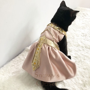 May include: A black cat wearing a pink and gold dress with a jeweled design. The dress has a gold belt and a gold and jeweled trim. The cat is sitting on a white fluffy surface. The text "Snowy's Closet" is visible on the dress.