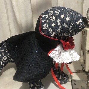 Puede incluir: Un perro de peluche negro y azul que lleva una capa negra con ribetes rojos y un collar de encaje blanco. El perro lleva una cinta roja alrededor del cuello. El perro est&aacute; sentado sobre una superficie blanca.