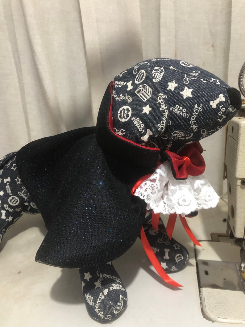 Puede incluir: Un perro de peluche negro y azul que lleva una capa negra con ribetes rojos y un collar de encaje blanco. El perro lleva una cinta roja alrededor del cuello. El perro est&aacute; sentado sobre una superficie blanca.