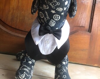 Puede incluir: Un juguete de peluche de perro con un esmoquin negro, camisa blanca y pajarita negra. El perro está sentado en un suelo de baldosas. El pelo del perro es azul con estrellas blancas y otros diseños.