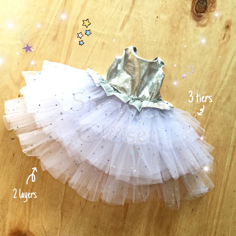 May include: A white tulle tutu with silver glitter stars and a silver bodice. The tutu has three tiers and two layers of tulle.