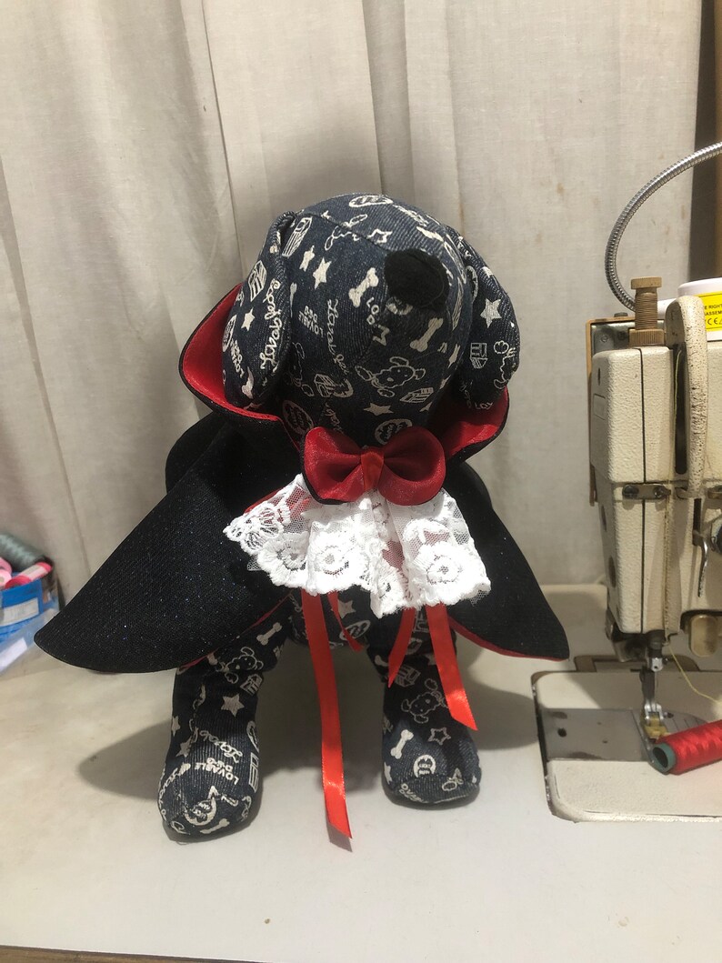 Puede incluir: Un perro de peluche negro y rojo vestido de vampiro para Halloween. El perro lleva una capa negra con forro rojo, un collar de encaje blanco y una pajarita roja. El cuerpo del perro est&aacute; cubierto de un patr&oacute;n blanco y negro con estrellas, huesos y las palabras "Love you lots".