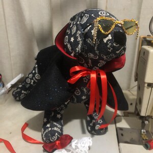 Puede incluir: Un perro de peluche negro y azul con una capa roja y gafas de sol brillantes. El perro tiene una cinta roja alrededor del cuello y sostiene un encaje blanco.