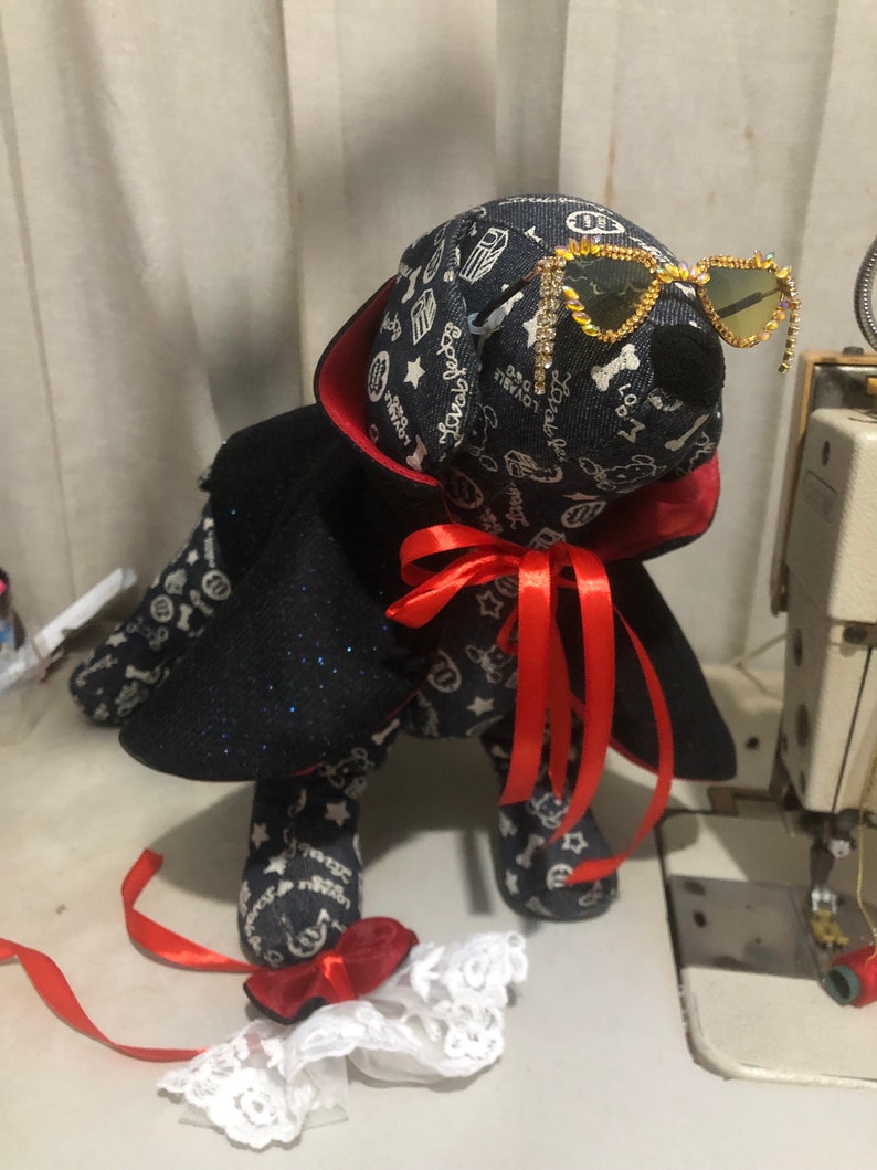 Puede incluir: Un perro de peluche negro y azul con una capa roja y gafas de sol brillantes. El perro tiene una cinta roja alrededor del cuello y sostiene un encaje blanco.