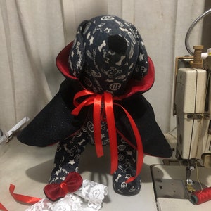 Puede incluir: Un perro de peluche negro y rojo vestido de vampiro con una capa negra, un collar rojo y una cinta roja. El perro lleva un brazalete de encaje blanco con un lazo rojo en cada pierna.